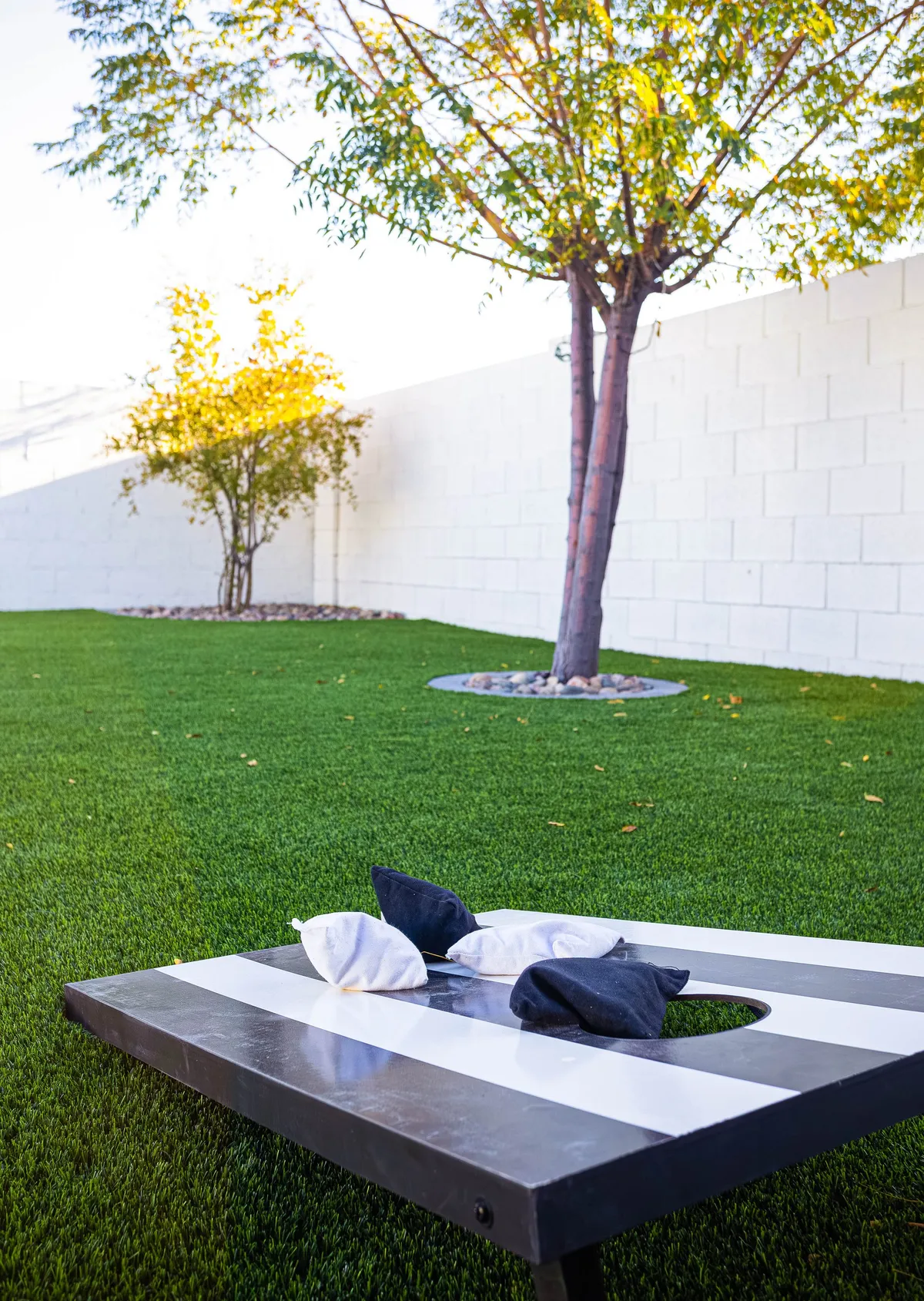 A spacious green lawn area dotted with trees, featuring a cornhole game board set up on the grass. The setting is outdoors and feels playful and relaxed, encouraging guests to enjoy lawn games and outdoor activities in the fresh air.