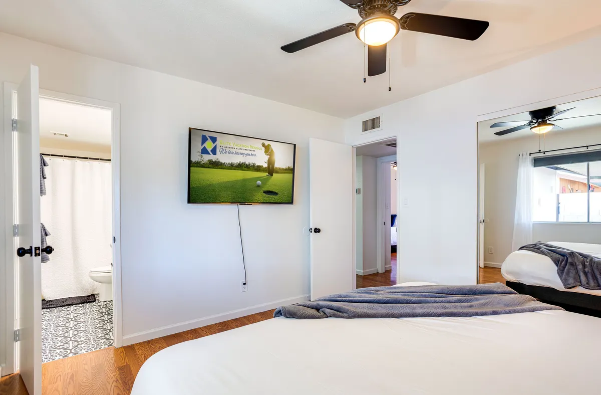 A comfortable guest bedroom featuring a large bed, plush linens, and a wall-mounted smart TV with a golf game on screen. The room is well-lit and includes a bathroom door, offering a glimpse of how bedrooms double as personal retreats for unwinding and entertainment.