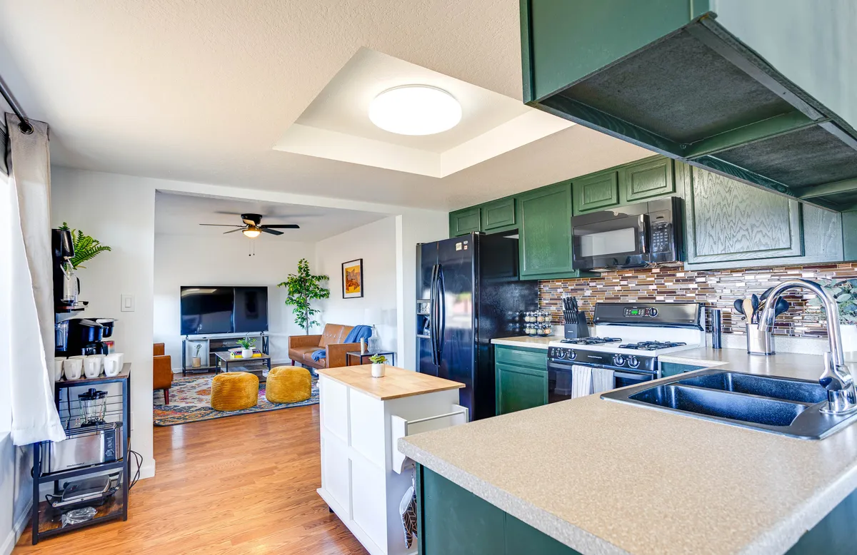 The image highlights an open-concept kitchen and living area, with a kitchen island, green cabinets, and a clear view into the welcoming living room. There’s evidence of shared activities and gatherings, blending the kitchen seamlessly with the relaxation space.