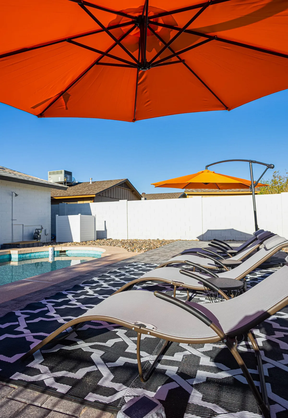 A spacious outdoor pool area with vibrant orange umbrellas, comfortable lounge chairs, and a textured outdoor rug. The setting is inviting, perfect for sunbathing, swimming, or socializing against a backdrop that hints at fun group gatherings and relaxation.