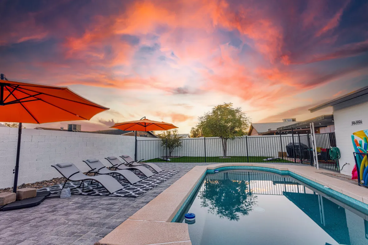 This image features a vibrant backyard scene with a sparkling swimming pool, surrounded by orange umbrellas and lounge chairs. Colorful pool toys are scattered on the green grass, and the area exudes a lively, sun-soaked atmosphere perfect for both relaxation and play.
