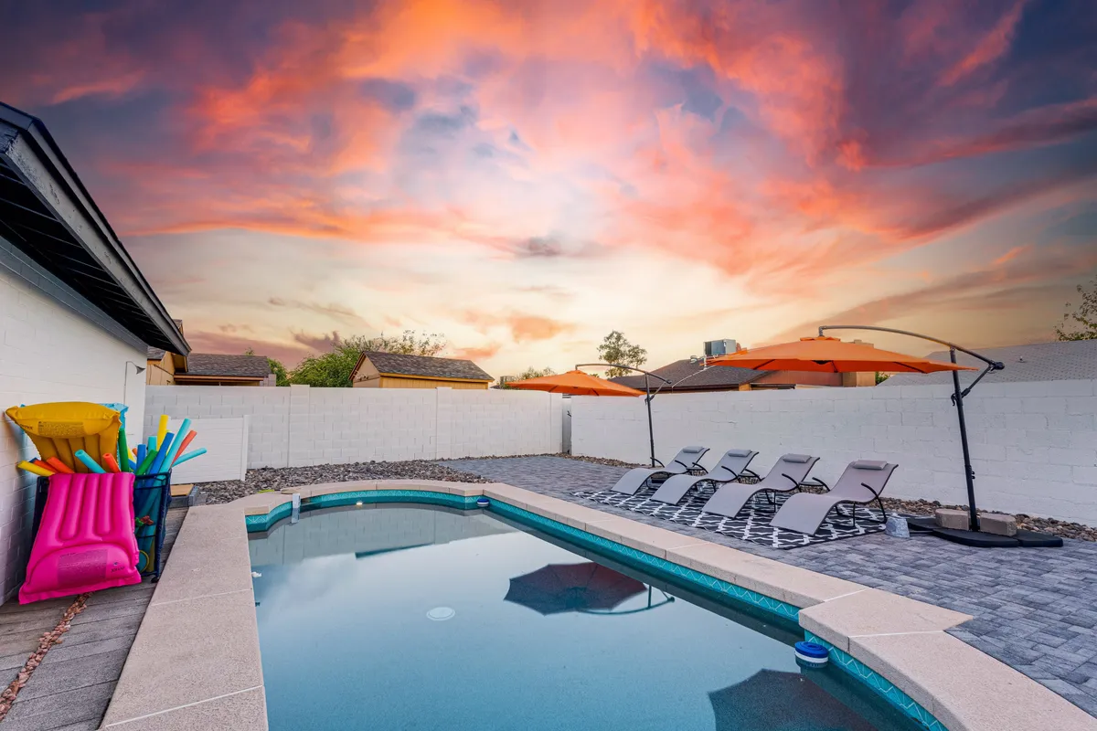 A vibrant backyard scene featuring a sparkling swimming pool surrounded by orange umbrellas and lounge chairs, with colorful pool toys floating in the water. The area is bathed in sunlight, suggesting an energetic and fun atmosphere ideal for both relaxation and play. The presence of seating and shade invites gatherings and socializing.