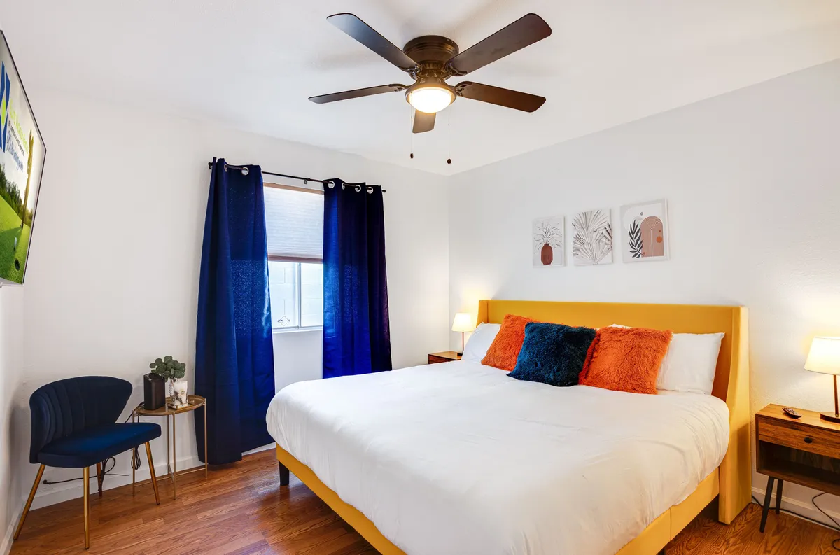 This image features a bedroom with a yellow bed frame, colorful pillows, blue curtains, a ceiling fan, and lively wall art. The overall atmosphere is inviting and serene, embodying the comfort and restful luxury described for the King Primary Suite. The soft bedding and cozy reading nook by the window reinforce the idea of a private retreat.
