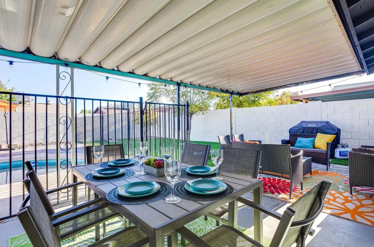 This exterior shot captures a backyard dining table set for a meal, with the swimming pool and colorful outdoor rug in the background. Comfortable seating creates a welcoming space for al fresco dining and gatherings.