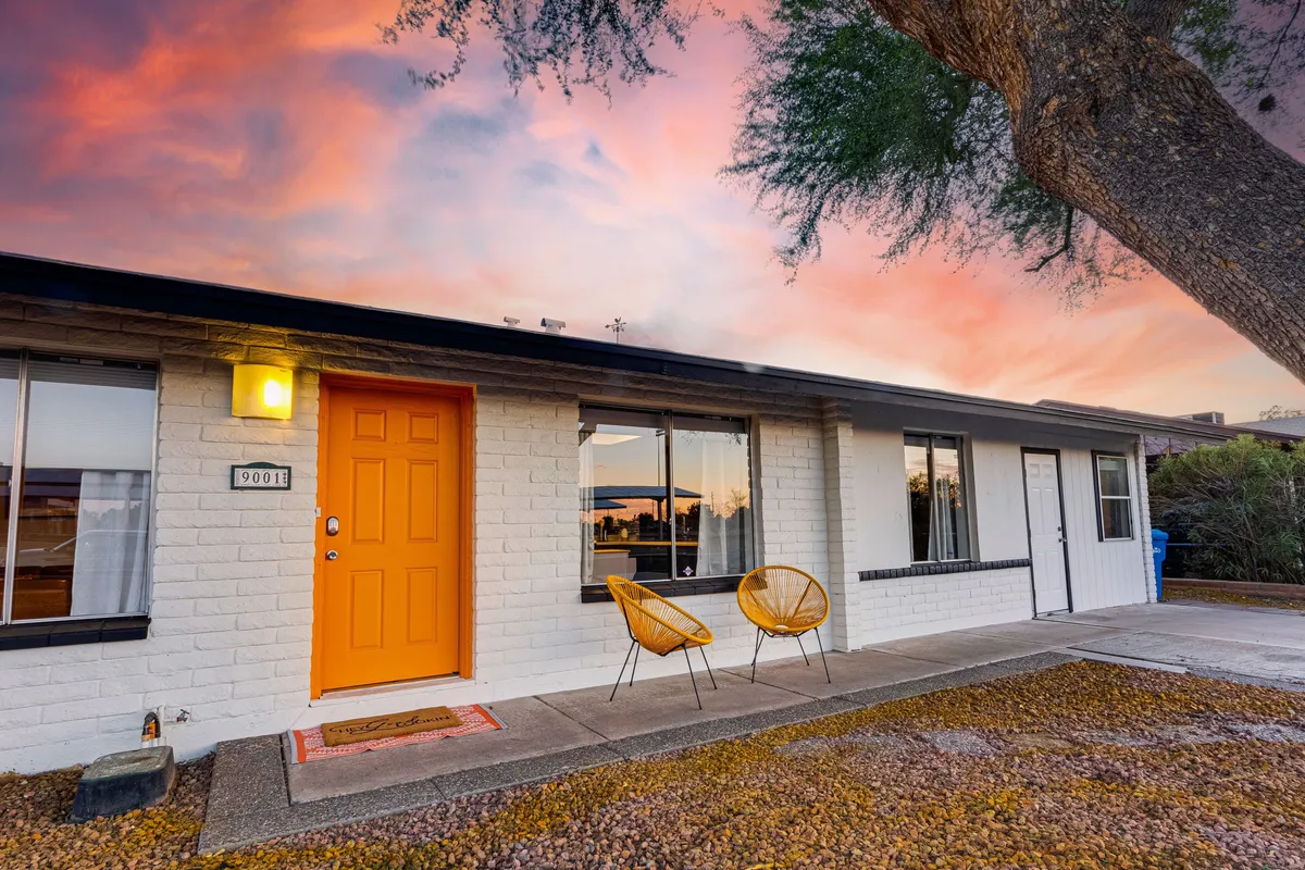 A vibrant exterior shot of the home's entrance, showcasing a striking orange door, stylish outdoor chairs, and a colorful sunset sky. The atmosphere is inviting and lively, ideal for welcoming groups back after big events or nights out.