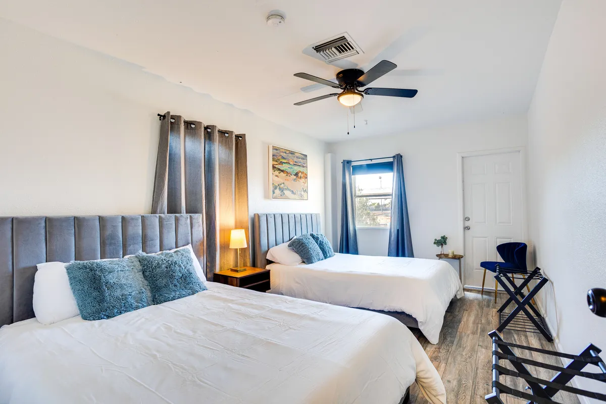 This image depicts a guest bedroom with two beds, a nightstand with a lamp, cheerful artwork, and a window with light curtains. The mood is comfortable and relaxing, with ample natural light and thoughtfully arranged beds and linens.
