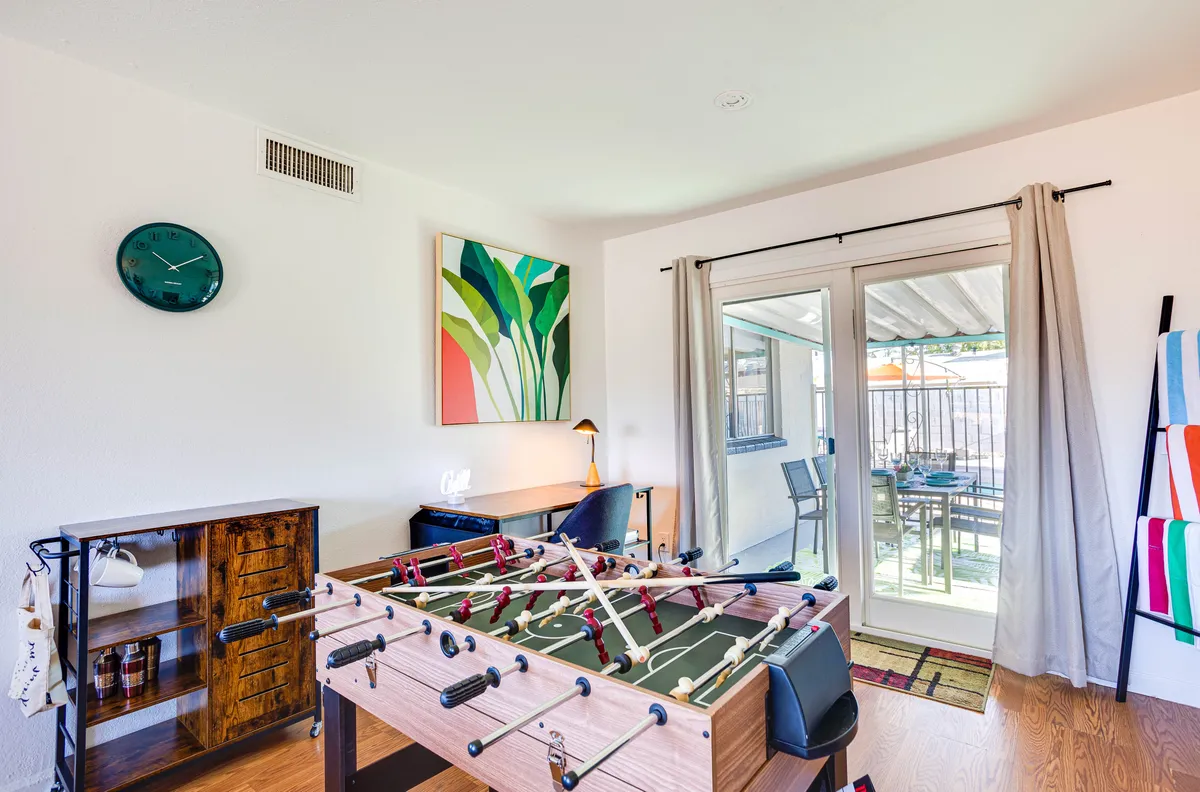This image showcases a lively work/game area with a foosball table set in the foreground, a large piece of artwork on the wall, a desk with a lamp for remote work or study, and a sliding glass door providing natural light. The arrangement offers a clear view of both the play and productivity zones, suggesting a space designed for both fun and focused tasks.